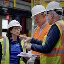Three team members in construction safety gear stand talking inside the large warehouse where materials testing is taking place.